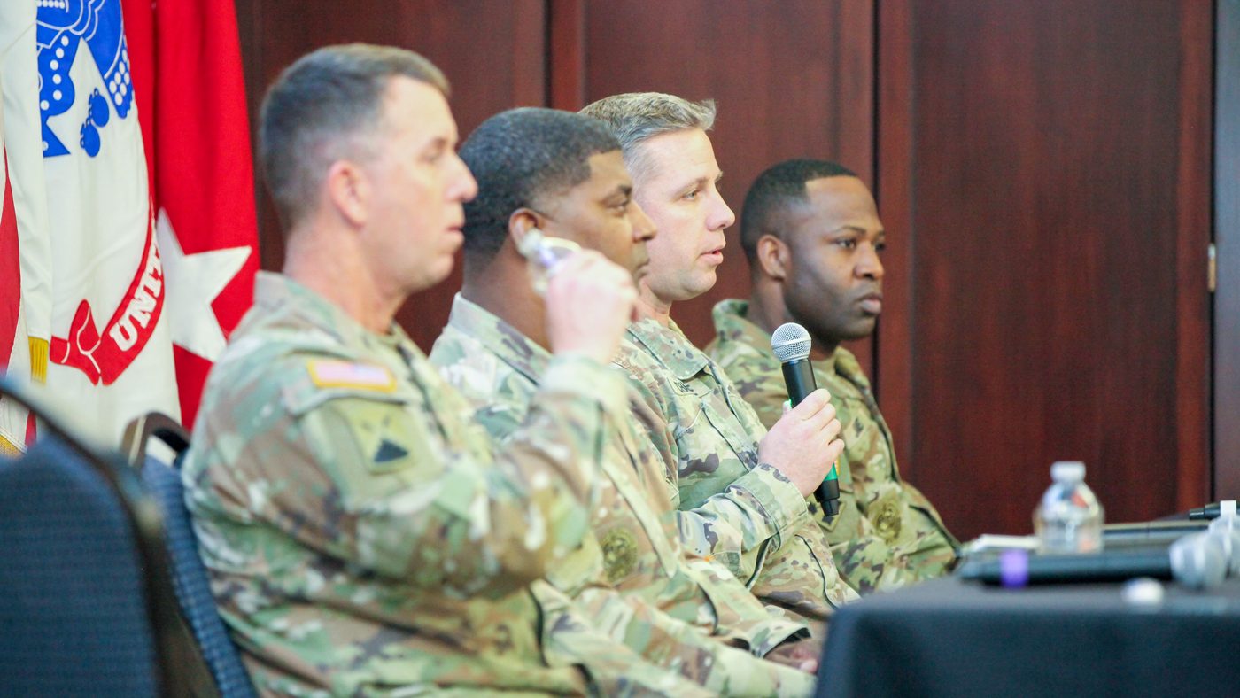 Uniformed panelists seated on stage, one speaking into a microphone, at a chapter program